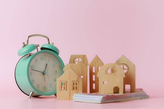 A bell alarm clock with wooden house model and banknotes on pink background for the concept of time management, finance, investment, housing and property.