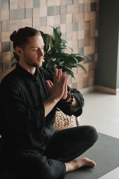 Portrait of a young man in a black kimano sitting in a lotus position on a gym mat in the interior.