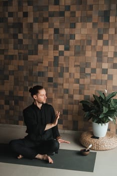 Portrait of a young man in a black kimano sitting in a lotus position on a gym mat in the interior.