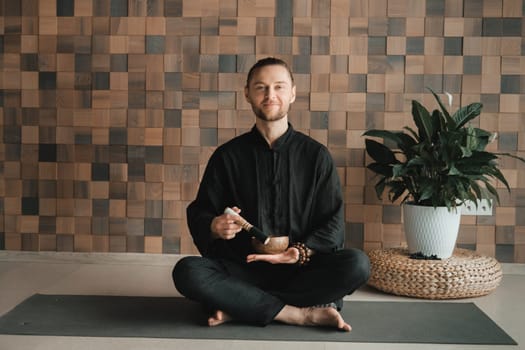 Portrait of a young man in a black kimono, holding a Tibetan bowl in his hands, sitting in a lotus position on a gym table in the interior.