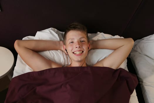 A handsome laughing boy lies on the bed with his head on the pillow and put his hands under his head in headphones. Boy teenager covered with a red blanket.