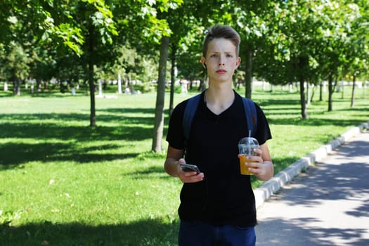 A handsome and smart boy of 14-17 years old holds a smartphone and a plastic glass with a straw with a soft drink in his hands. A young man in a black T-shirt and blue jeans looks at the camera in a leisure park.