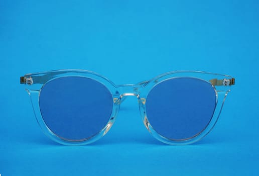 Sunglasses with a transparent frame and blue lenses on a blue paper background. Plastic glasses front view.