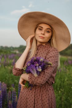 A beautiful woman in a straw hat walks in a field with purple flowers. A walk in nature in the lupin field.