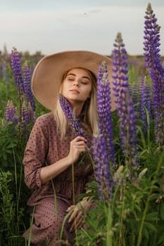 A beautiful woman in a straw hat walks in a field with purple flowers. A walk in nature in the lupin field.