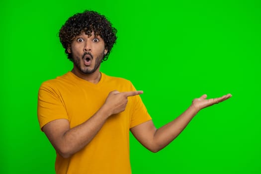 Young indian man showing thumbs up and pointing empty place, advertising area for commercial text, copy space for goods promotion. Bearded hindu guy isolated on green chroma key background, indoors