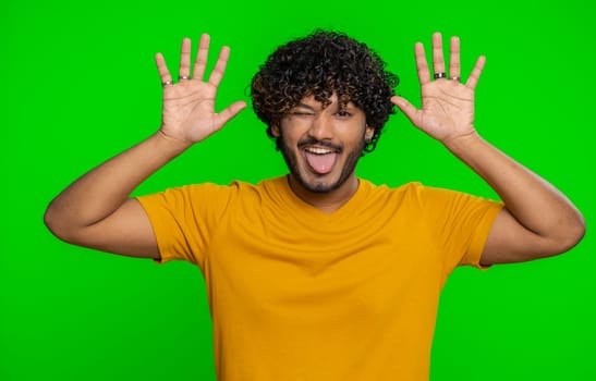 Comical funny young indian man making playful silly facial expressions and grimacing, fooling around, showing tongue. Handsome hindu crazy guy bullying isolated on chroma key background indoors