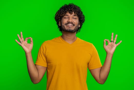 Keep calm down, relax, inner balance. Young indian man breathes deeply with mudra gesture, eyes closed, meditating with concentrated thoughts peaceful mind. Hindu guy isolated on chroma key background