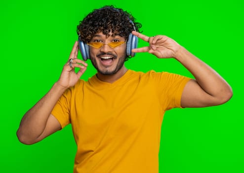Happy indian man in headphones listening music and dancing disco fooling around having fun expressive gesticulating hands, relaxing on party. Hindu guy isolated alone on studio chroma key background