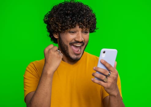 Happy excited joyful indian man in green shirt use smartphone typing browsing shouting say wow yes found out great big win, good news, lottery goal achievemen, celebrating success, winning game