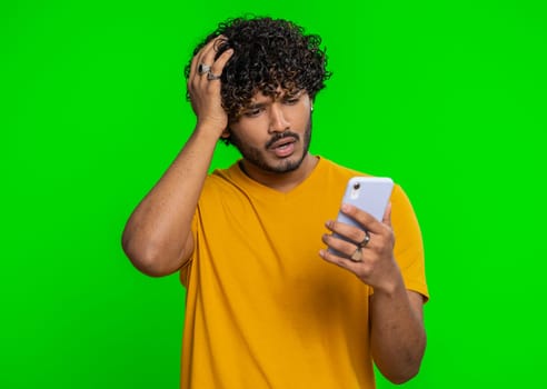 Indian man use mobile cell phone typing browsing, loses becoming surprised sudden lottery results, bad fortune, loss, fail. Handsome hindu guy isolated alone on green chroma key background, indoors