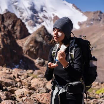 Beautiful young sportive girl applies sunscreen to her hands and face while climbing the mountain, woman takes care of her skin while traveling in sunny weather.