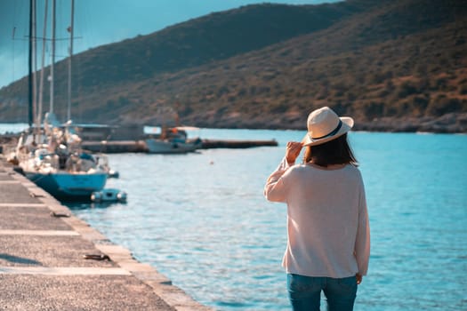 A young girl in a hat looks at the picturesque view with the sea and moored yachts on a sunny day, a backpacker travels and discoveries beautiful places on the coast during her holidays.