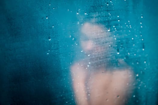 A young girl takes a shower, in the foreground there are drops on the misted glass, the woman is blurred.