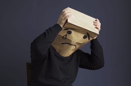 A sad man in a black turtleneck with a bag on his head, with a drawn crying emoticon, holds his head and cries. Emotions and gestures.
