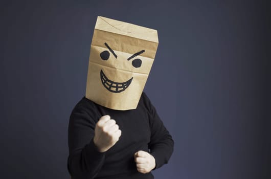 A man in a black turtleneck with a bag on his head, with a drawn angry emoticon, shows his fist while sitting on a chair. Emotions and gestures.