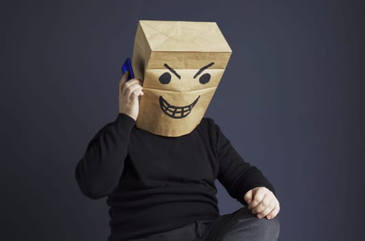 A man in a black turtleneck with a bag with an evil smiley on his head is talking on the phone while sitting on a chair. Emotions and anger