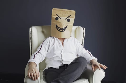 A man with a paper bag on his head, with a drawn angry smiley face, sits in a white chair. Emotions and gestures.