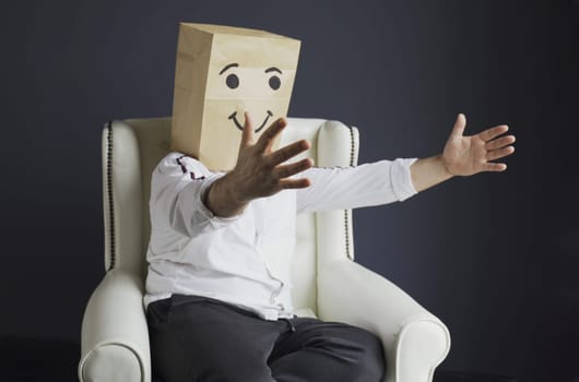 A man in a white shirt with a paper bag on his head, with a painted smiley, sits in an armchair, overwhelmed with feelings, stretches his arms for hugs.