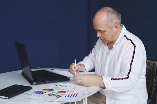 A male businessman works on a laptop, studies reporting documents, charts and graphs, makes notes in a notebook.