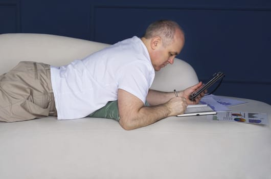 A businessman lies on a sofa and looks at data trends on charts and graphs in a tablet, makes notes in a notebook. Distant work