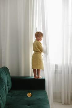 A boy in a yellow coat is standing on a green sofa near the window, next to it is half an orange.