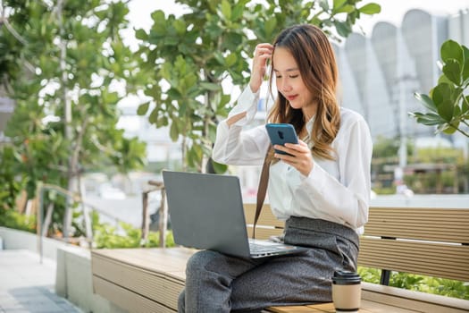 Asian business young woman working laptop and using mobile smartphone outdoor building exterior, Happy smiling businesswoman sit writing text on mobile phone outside street city near office in morning