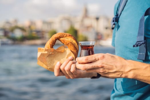 Man in Istanbul having breakfast with Simit and a glass of Turkish tea. Glass of Turkish tea and bagel Simit against golden horn bay and the Galata Tower in Istanbul, Turkey. Turkiye.