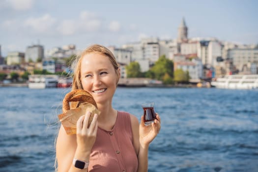 Woman in Istanbul having breakfast with Simit and a glass of Turkish tea. Glass of Turkish tea and bagel Simit against golden horn bay and the Galata Tower in Istanbul, Turkey. Turkiye.