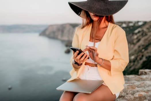 Successful business woman in yellow hat working on laptop by the sea. Pretty lady typing on computer at summer day outdoors. Freelance, travel and holidays concept.