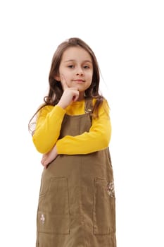Vertical studio portrait of 5-6 years Caucasian adorable kid girl in khaki and yellow dress, holding finger on her chin and reasoning seriously, looking at camera, isolated on white background.