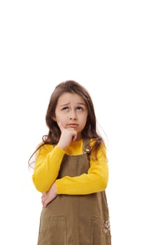 Thoughtful pensive little girl wearing casual khaki dress, holding chin, looking up, thinking about future and reasoning over problems, posing on white background with serious facial expression.