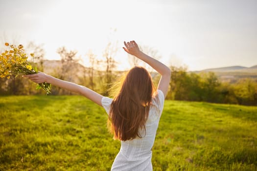 photo from the back of a red-haired woman raising her hands in happiness with a bouquet of flowers in the rays of the setting sun. High quality photo