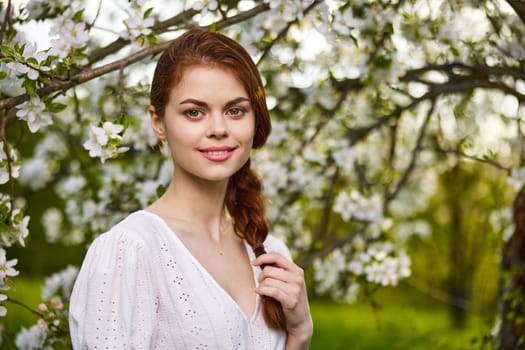 portrait of a joyful woman in a light dress against the background of a flowering tree, holding herself by her braid. High quality photo