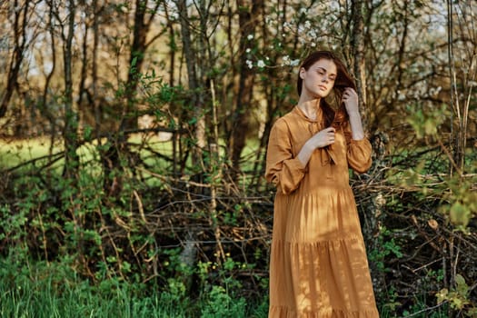 a beautiful woman walks in the shade near the trees, dressed in a long orange dress, enjoying the weather and the weekend, touching her long hair. The theme of privacy with nature, horizontal photography . High quality photo