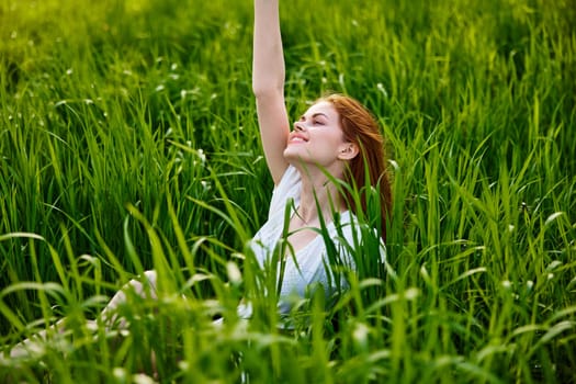 a cute woman in the summer high grass sits in a light dress happily raising her hand up. High quality photo