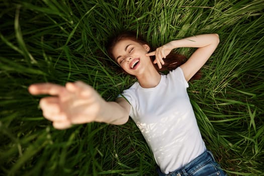 A happy girl in a white t-shirt stretches her arms up and lies on the green grass. High quality photo