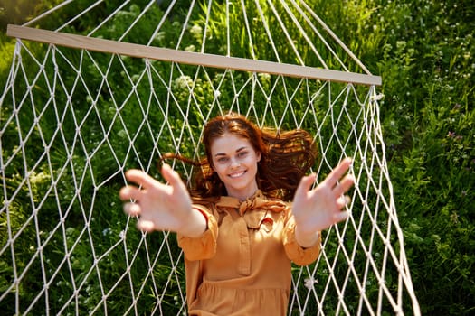 a beautiful, elegant woman lies in a long orange dress on a mesh hammock resting in nature, illuminated by the warm sunset light, smiling happily, stretching out her hands to the camera. High quality photo