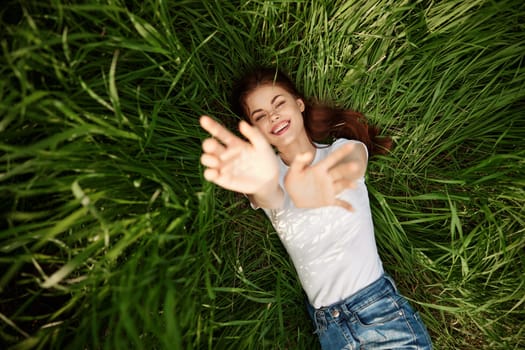 A happy girl in a white t-shirt stretches her arms up and lies on the green grass. High quality photo