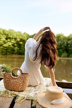 close-up photo of a woman sitting with her back to the camera on the shore of a lake with a wicker bag full of daisies. High quality photo