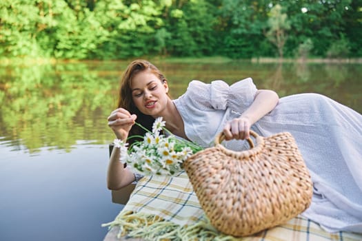 a lovely woman in a light dress lies on a pier by the lake on a sunny day with a basket of daisies in her hands. High quality photo