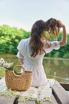 a woman in a light dress sits on the shore of the lake with her back to the camera and straightens her red hair. High quality photo