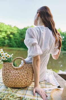 a woman in a light dress sits on the shore of the lake with her back to the camera and straightens her red hair. High quality photo