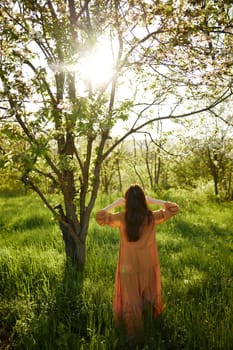 beautiful, a woman stands in a long orange dress, in the countryside, near a flowering tree, during sunset, stands with her back to the camera, holding her hands loosely near her head. High quality photo