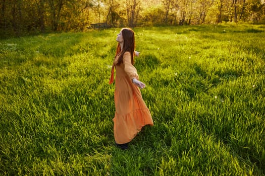 an attractive, slender, red-haired woman stands in a wide, green field during sunset in a long orange dress enjoying unity with nature and relaxation raising her arms to the sides while standing with her back to the camera. High quality photo