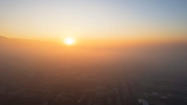 Epic gray smog is visible at sunset over the city. A bird's-eye view from a drone of houses, roads, cars and parks. White clouds and snowy mountains are illuminated by orange rays of the sun. Almaty