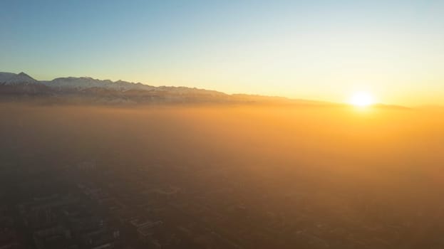 Epic gray smog is visible at sunset over the city. A bird's-eye view from a drone of houses, roads, cars and parks. White clouds and snowy mountains are illuminated by orange rays of the sun. Almaty