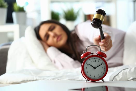 Young woman tries to smash an alarm clock with wooden mallet and destroy clock. Displeased angry girl lies in bed and turns off the alarm clock with hammer