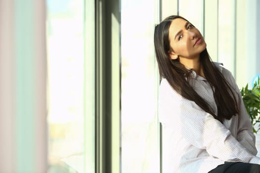 Dreamy young business woman is sitting on windowsill near huge window in modern office sun is shining brightly. Businesswoman enjoying break and relaxing after a hard day at work