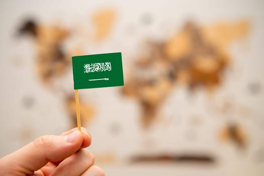 Saudi Arabia flag in mans hand on the wooden world map background. Global economy and geopolitics concept.